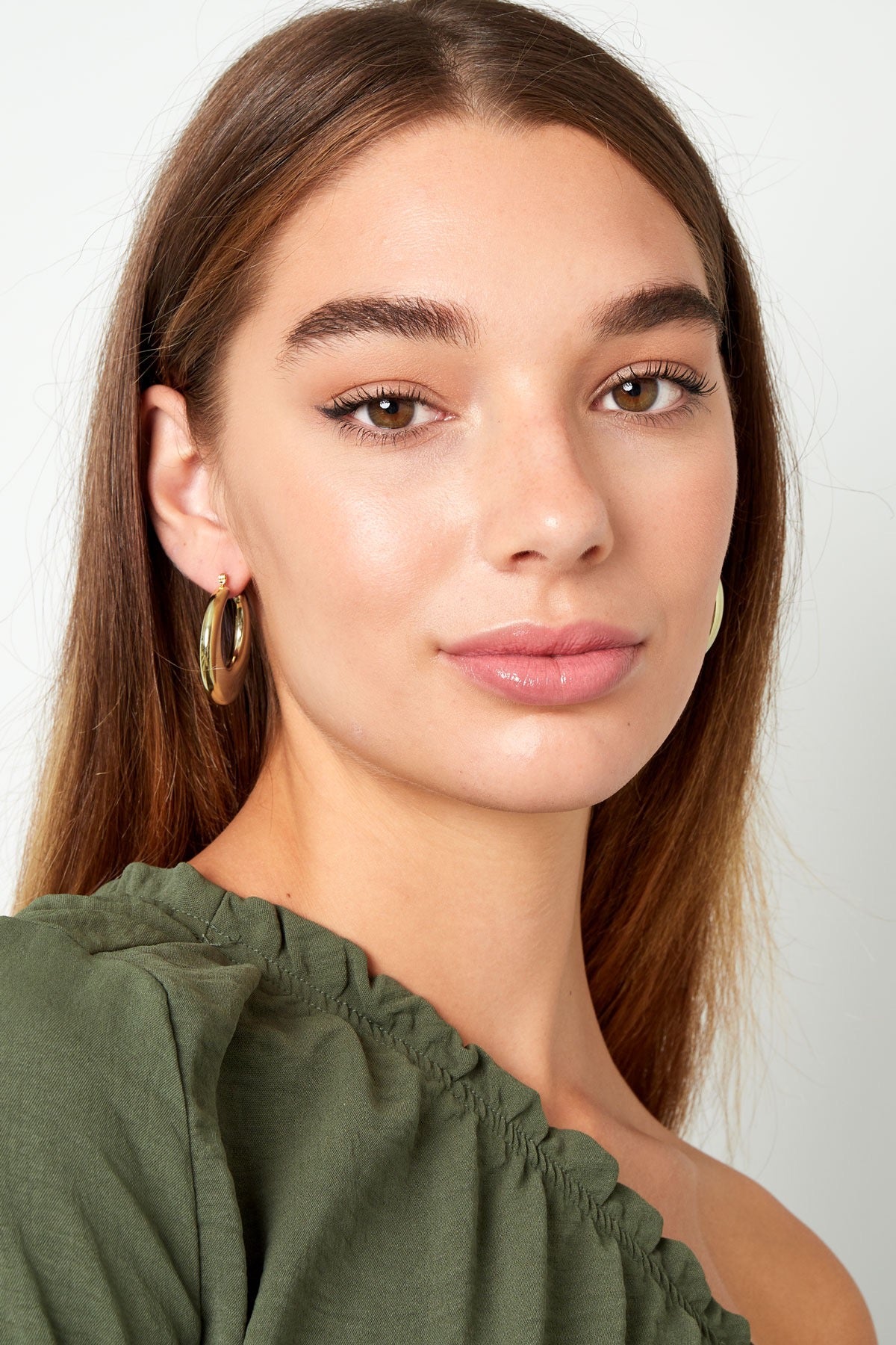Close-up van een vrouw met glanzende gouden Oorbellen | Rond van Louxx the brand, met een licht glimlach en casual groene top.