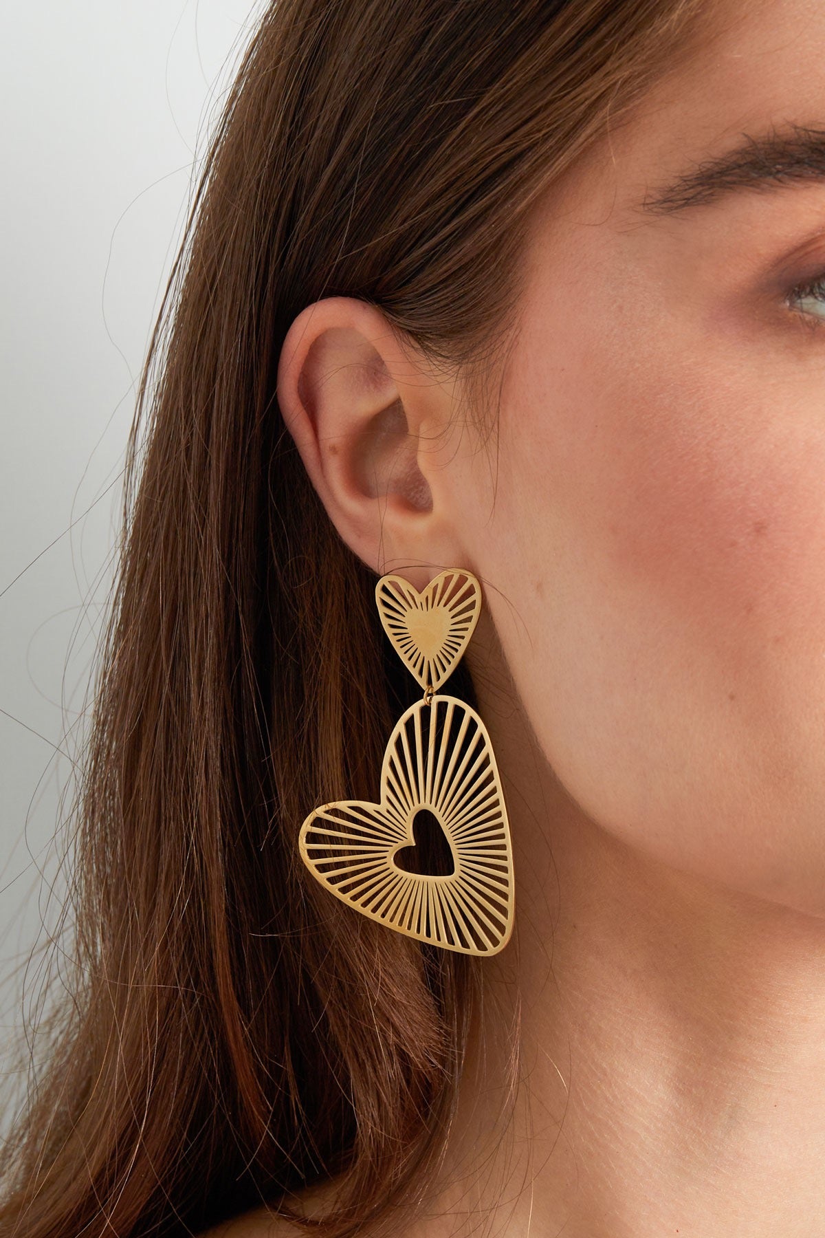 Statement gouden oorbellen met twee verbonden harten en een uitgesneden patroon, gedragen door een model met lange bruine haren.