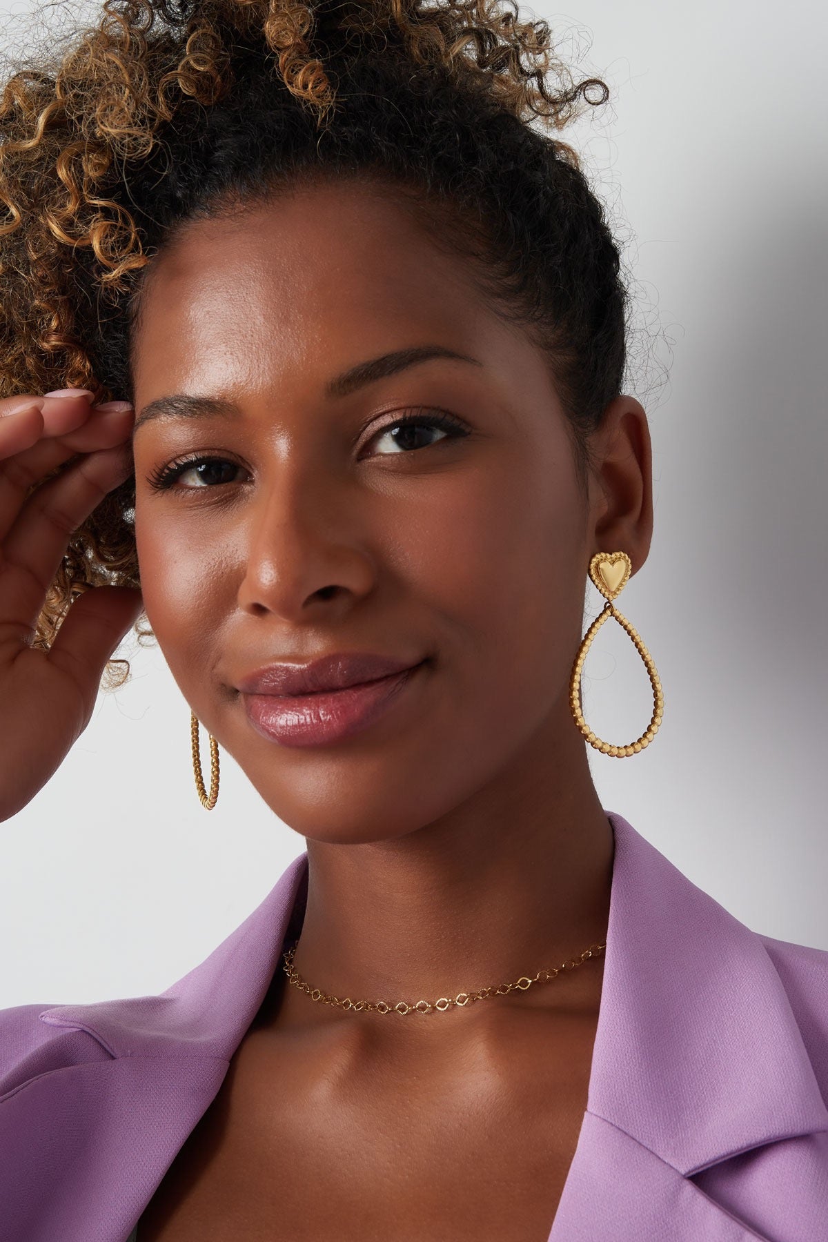 Vrouw die gouden hart oorbellen van Louxx the brand draagt. De oorbellen hebben een elegante druppelvorm en voegen een speelse touch toe aan haar outfit.