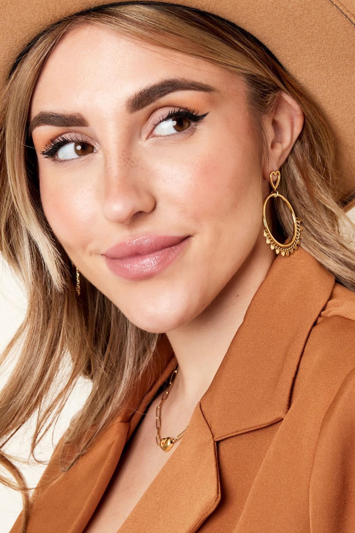 Een vrouw in een trendy outfit met gouden hart oorbellen van Louxx the brand. Haar haar en make-up zijn stijlvol, wat de elegantie van de oorbellen accentueert.