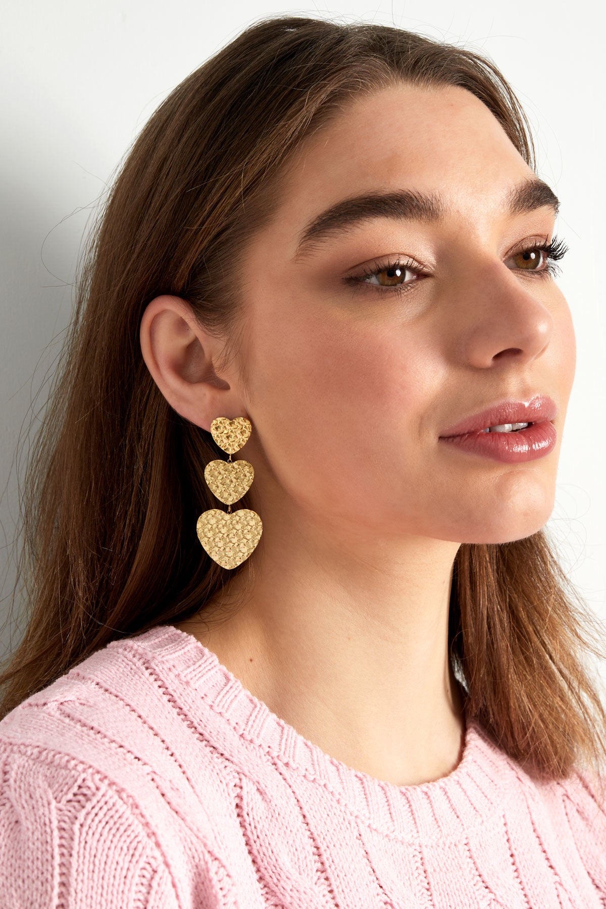 Model draagt gouden hart oorbellen van Louxx the brand, met drie hangende hartvormige elementen en een delicate uitstraling. De hart oorbellen goud zijn ideaal voor stijlvolle outfits.