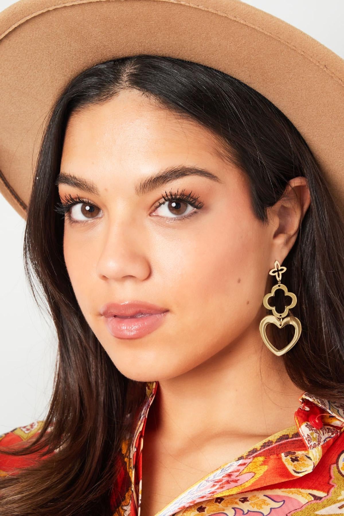 Close-up van een vrouw die gouden oorbellen met 3 bedels van Louxx the brand draagt. De oorbellen zijn elegant en passen goed bij haar kleurrijke blouse.